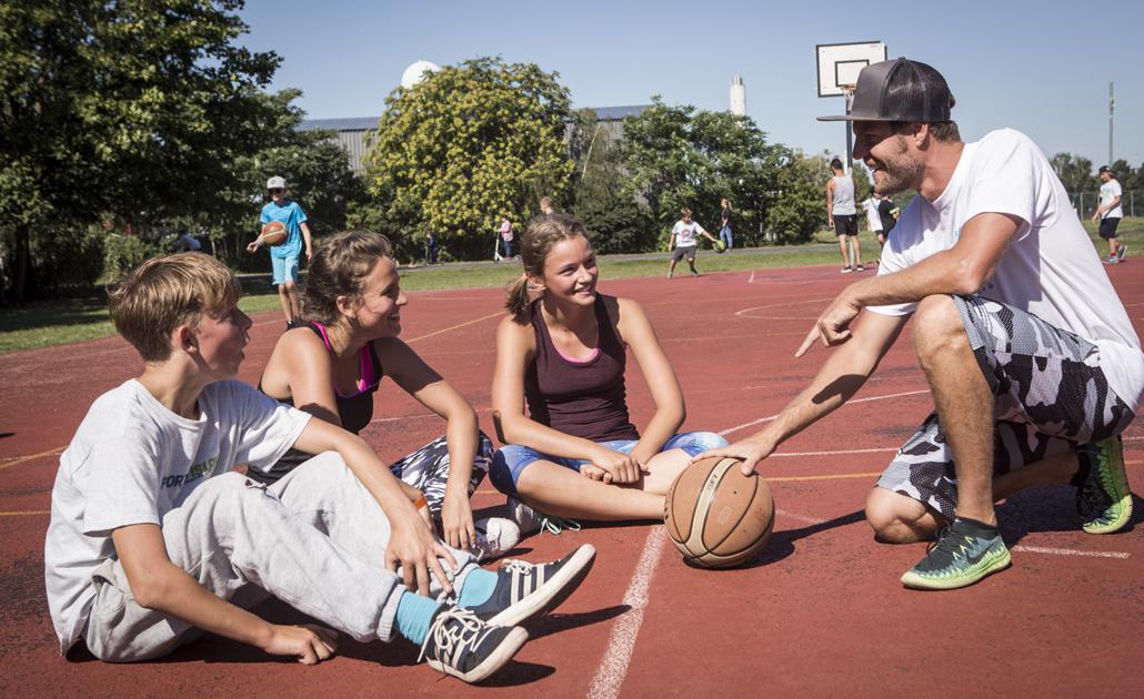 Sportpädagogen Arne sitzt mit Kindern auf dem Basketballplatz und erklärt Basketballregeln