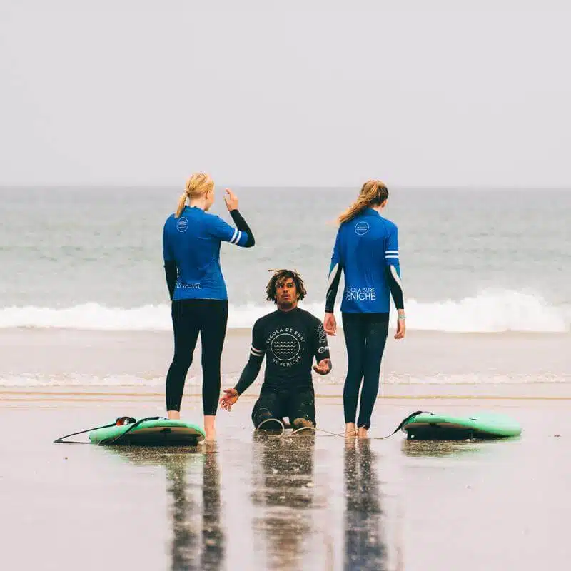 Teenager mit Coach stehen auf den Jugendreisen Portugal während des Surfkurses am Strand mit Wellen am Horizont